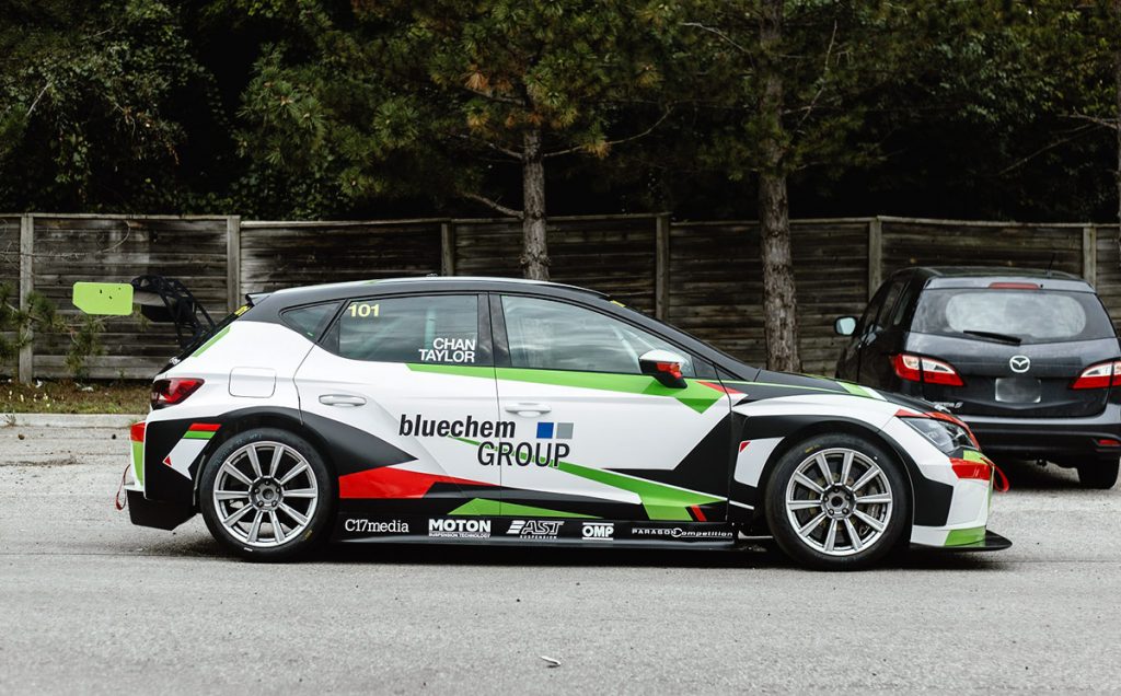 Race car featuring a custom bluechem GROUP livery, showcasing vibrant colors and sleek design, parked in a motorsport setting with trees and other vehicles in the background.