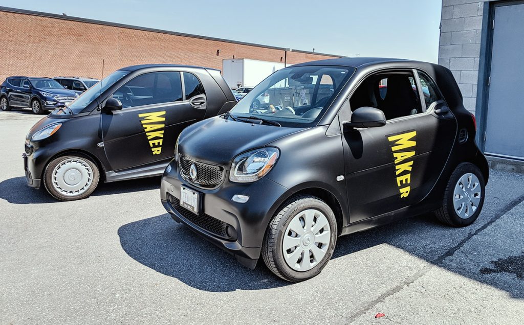 Two smart cars with bold black vehicle wraps displaying the word "MAKER," parked in a lot, showcasing effective commercial vehicle branding for advertising purposes.