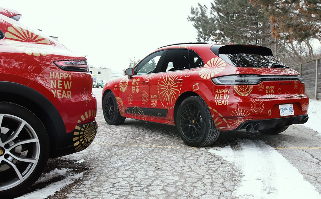 Red Porsche Macan with festive "Happy New Year" vehicle wrap, showcasing vibrant graphics and branding, parked in a snowy outdoor setting.