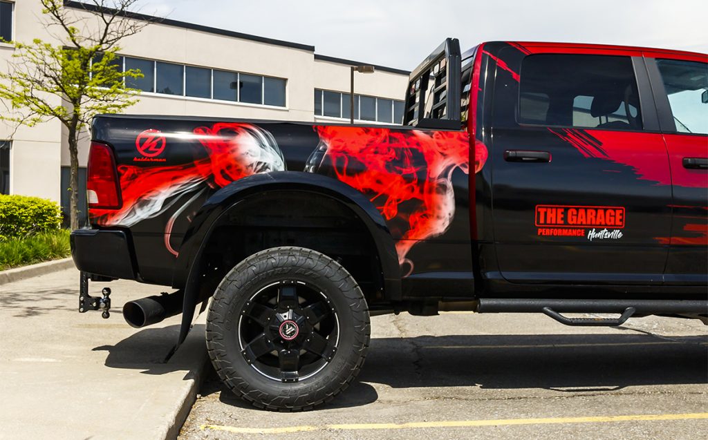 Commercial vehicle wrap featuring bold red and black design with "THE GARAGE PERFORMANCE" branding on a pickup truck, parked in a business setting with greenery and modern architecture in the background.