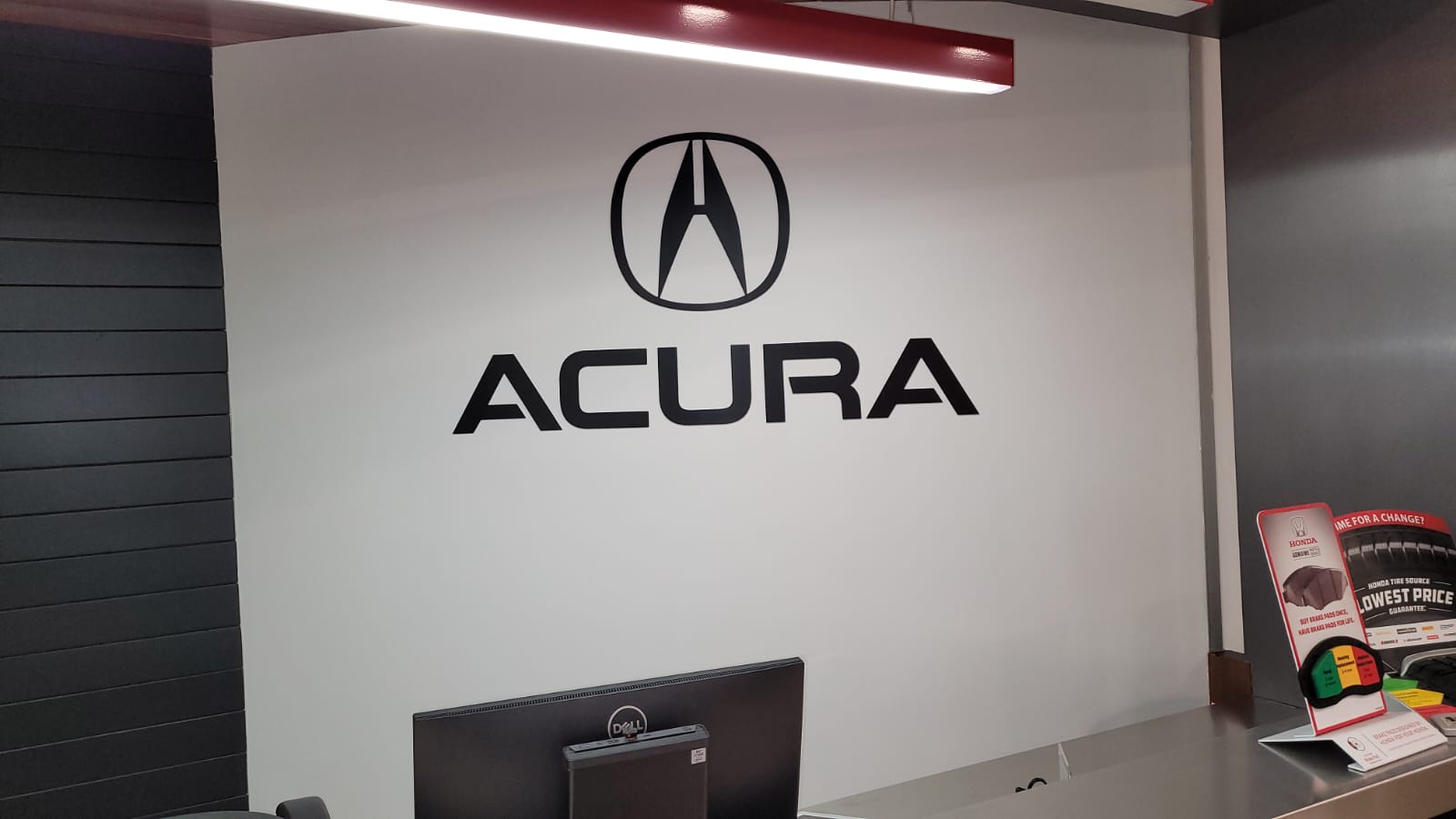 ACURA logo displayed prominently on a white wall in a modern setting, with a computer monitor and promotional materials visible in the foreground.